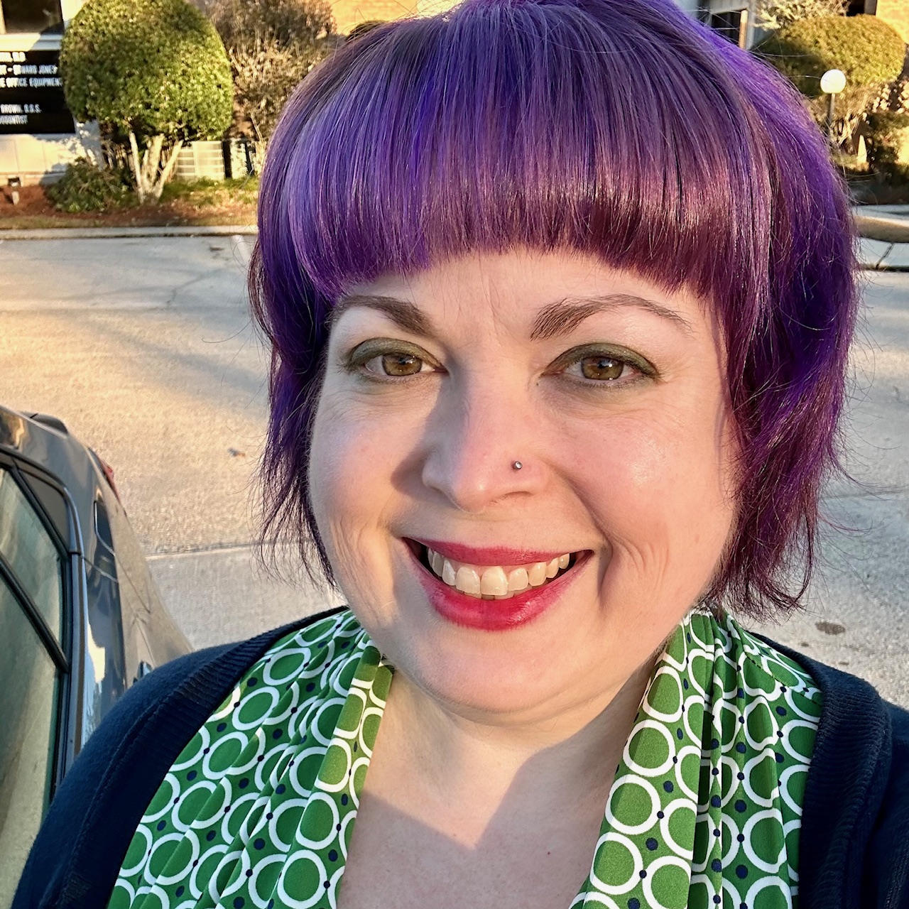 Picture of Karen. She has a purple bob hairstyle, is wearing a green patterned dress and a navy cardigan, and is smiling at the camera.