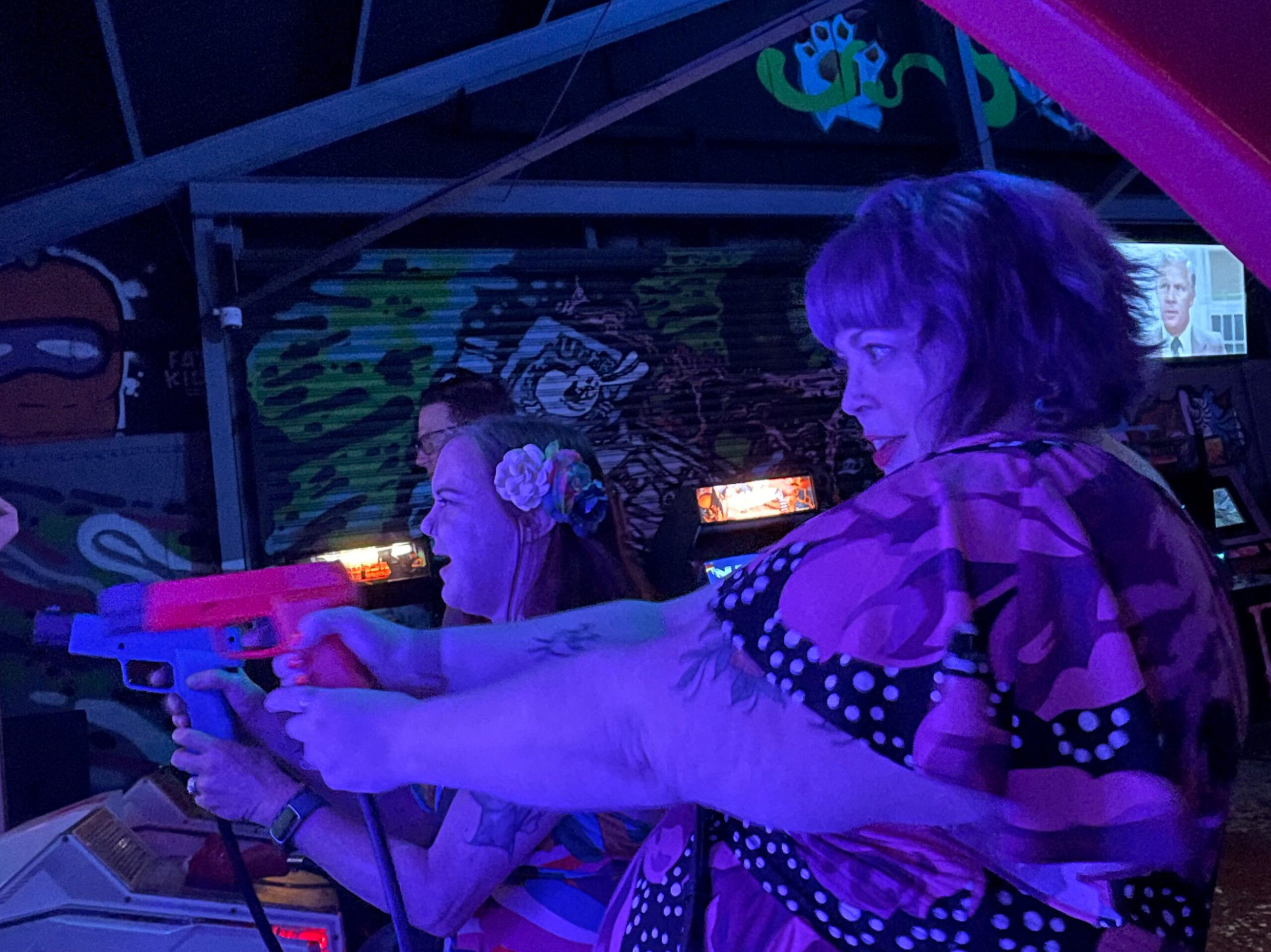 Two women bathed in blue light at a arcade, holding red and blue guns and playing a shooting game.
